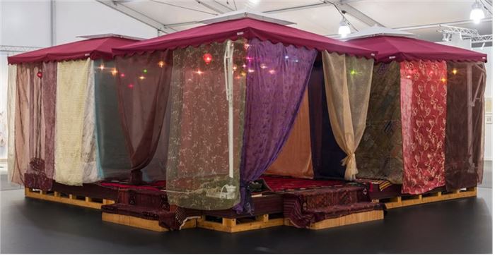 Chris Burden, Nomadic Folly, 2001, wood platform, 4 cloth and metal umbrellas, woven carpets, braided ropes, pillows, silken fabrics, glass and metal lamps, iPod, and speakers. Courtesy of Gagosian.